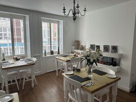 A dining area with a wooden table, white chairs, and a chandelier. There are windows with blinds, a fireplace, and a shelf with decorative items.
