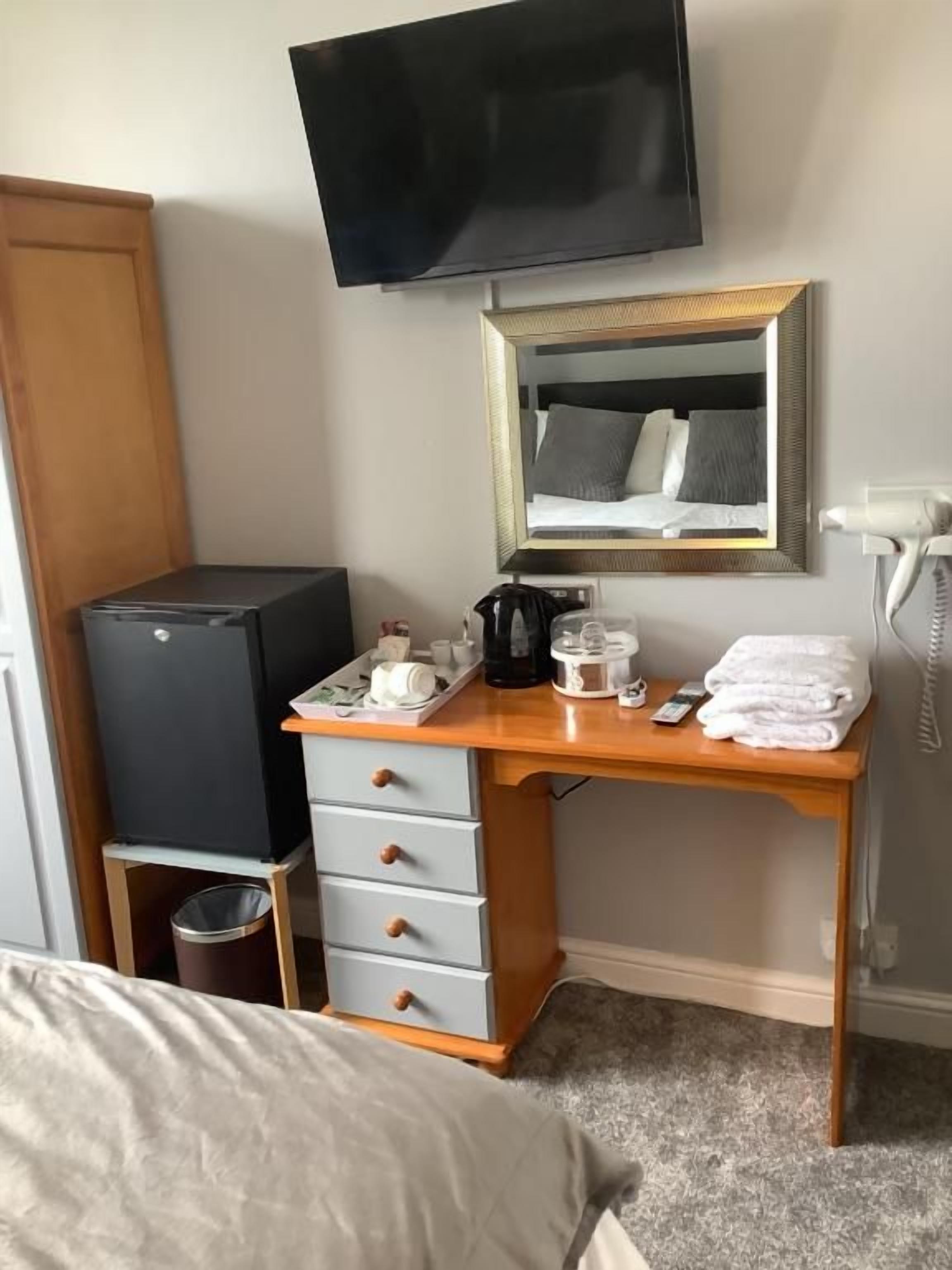 A hotel room with a bed, a desk with a mirror, a television mounted above the desk, a hairdryer, and a trash bin.