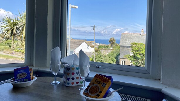 A table with a bowl of cereal, a box of cereal, and a glass of water by a window with a view of houses and the sea. - Beechwood House (St Ives)