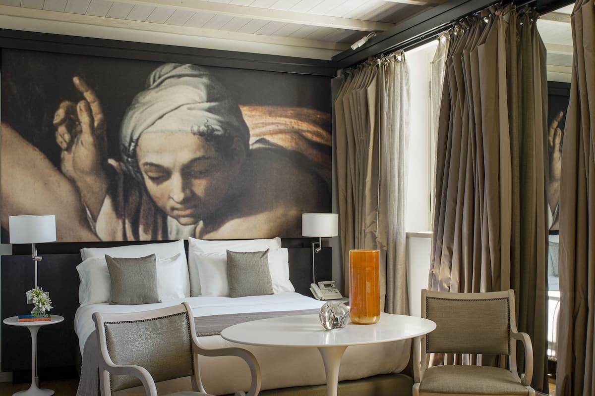 Una camera d'albergo con un letto, due sedie, un tavolino, una lampada e un quadro appeso al muro.