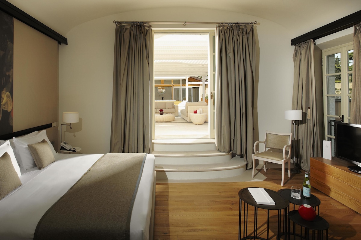 Habitación de hotel con una cama grande, un escritorio con una computadora, una silla y un televisor. Hay un cuadro en la pared y una ventana con cortinas.