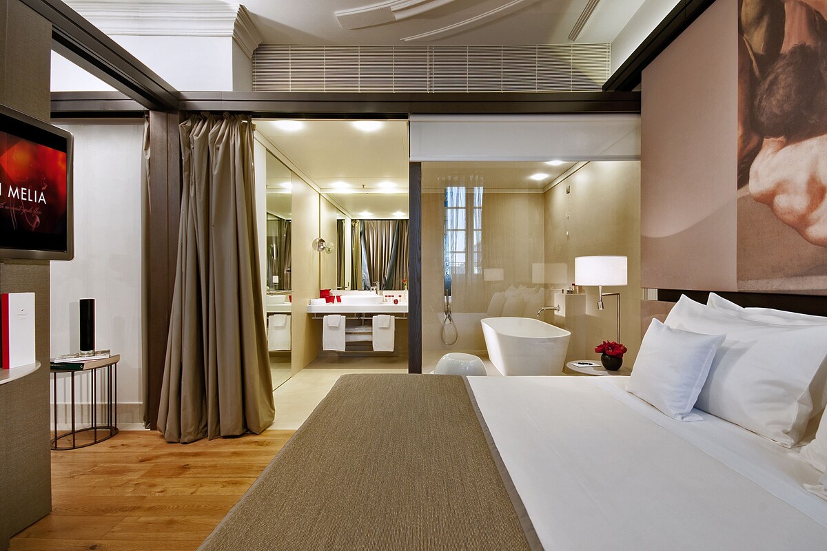 A hotel room with a bed, a bathroom, a TV, and a large window.