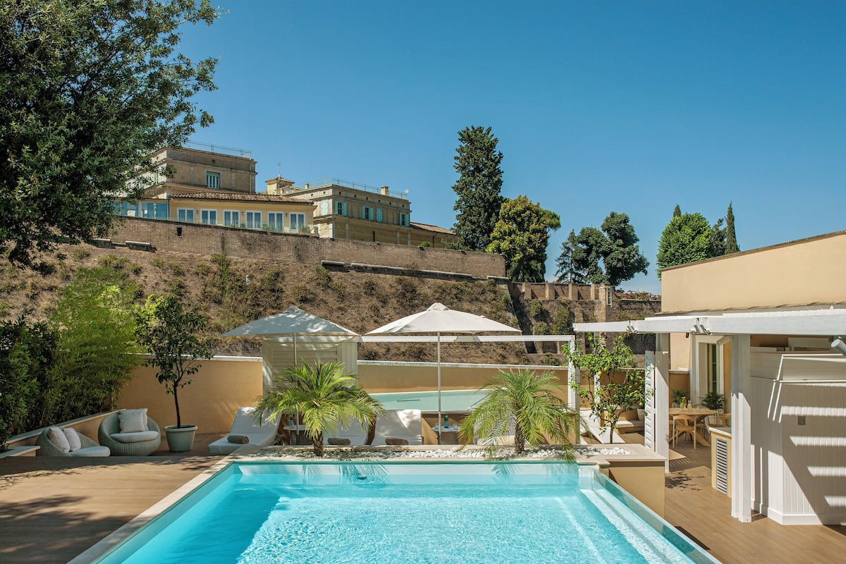 Una piscina con lettini e ombrelloni, circondata da un giardino curato e vista su una collina.
