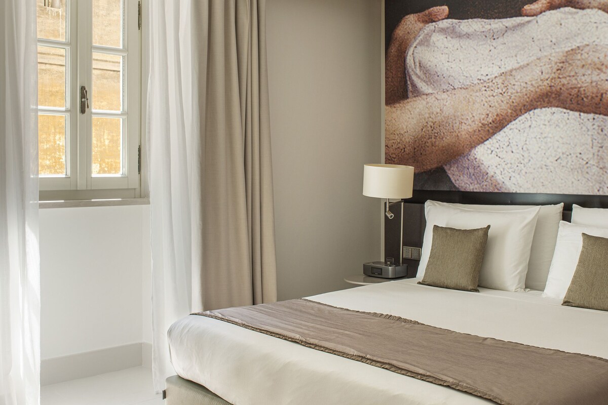 A bedroom with a bed, curtains, a painting, and a lamp.