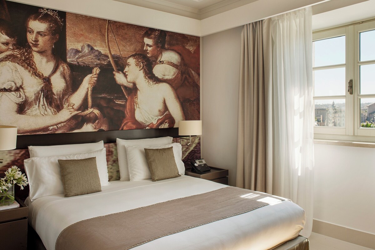 Habitación de hotel con un cuadro clásico grande en la pared, una cama bien hecha con ropa de cama blanca y marrón, una mesita de noche con lámpara y una ventana con cortinas.