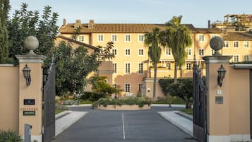A gated entrance to a building with a symmetrical facade, a central entrance, and a well-maintained garden with palm trees and shrubs.