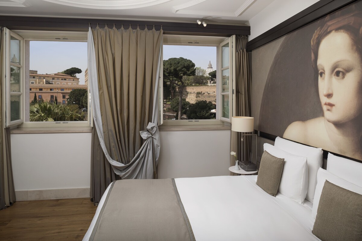 Habitación de hotel con una cama grande, una mesita de noche, una lámpara y vistas de la ciudad a través del ventanal.