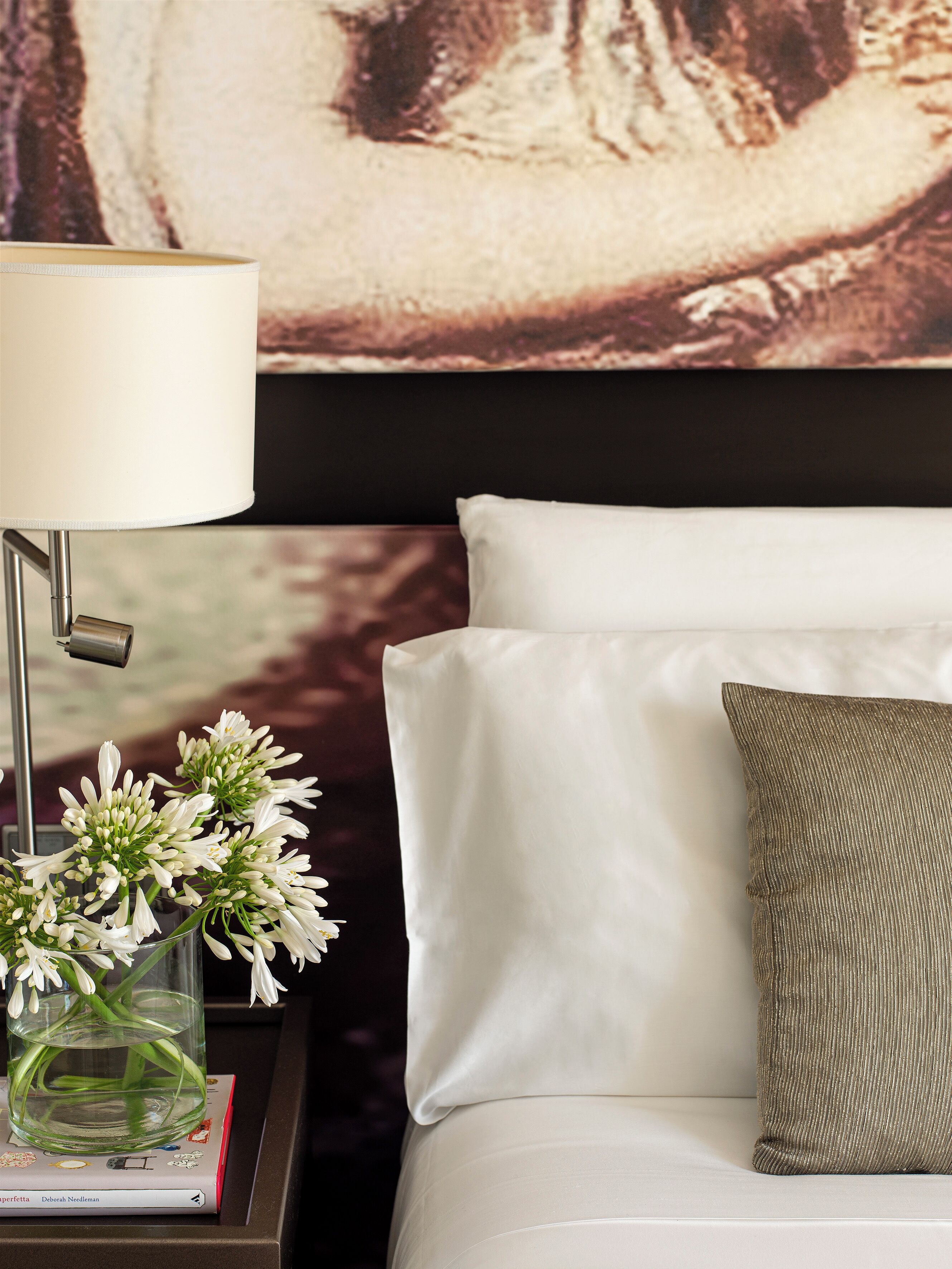 A neatly made bed with white linens, a green pillow, and a bedside table with a lamp and a vase of flowers.