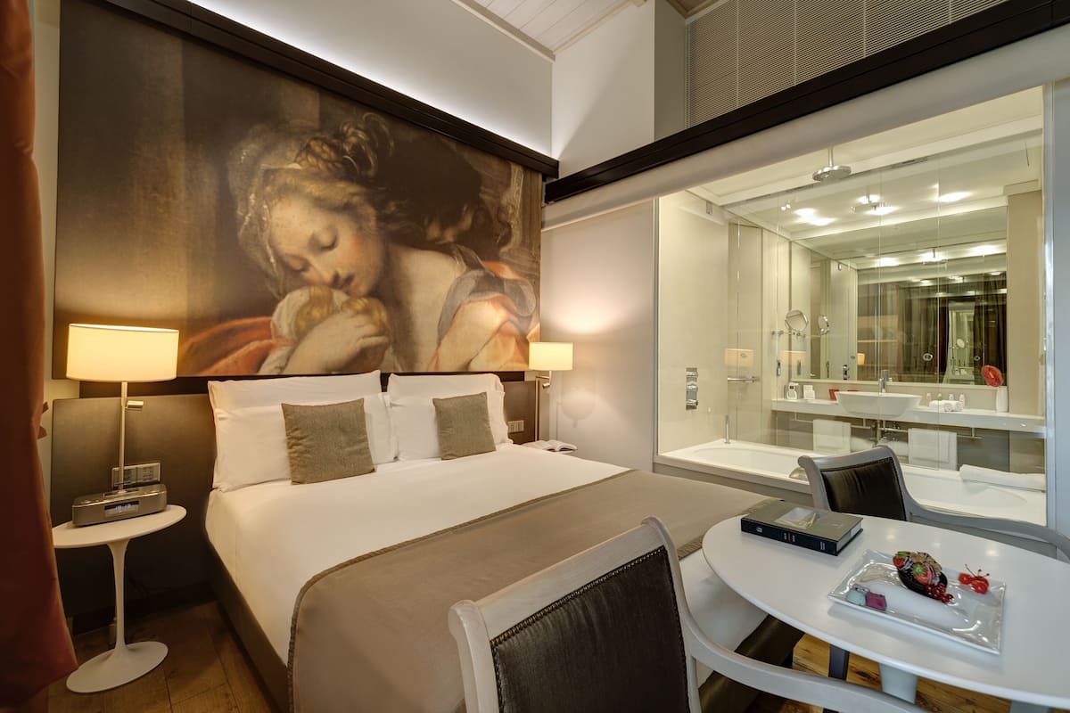 Camera d'albergo con un letto grande, lampade sui comodini, un tavolino con un vassoio di cibo, uno specchio grande e un quadro appeso al muro.