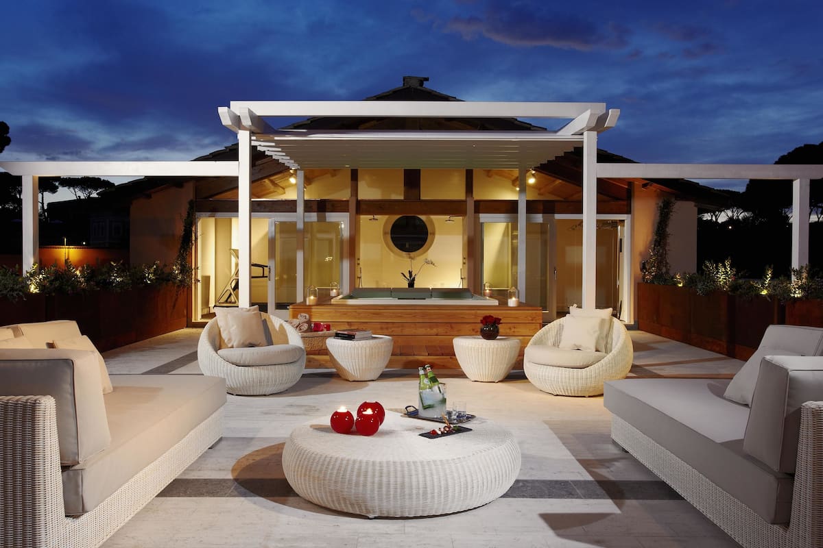 Una zona living esterna moderna con mobili in rattan, un'area salotto centrale e vista sulla casa sullo sfondo.