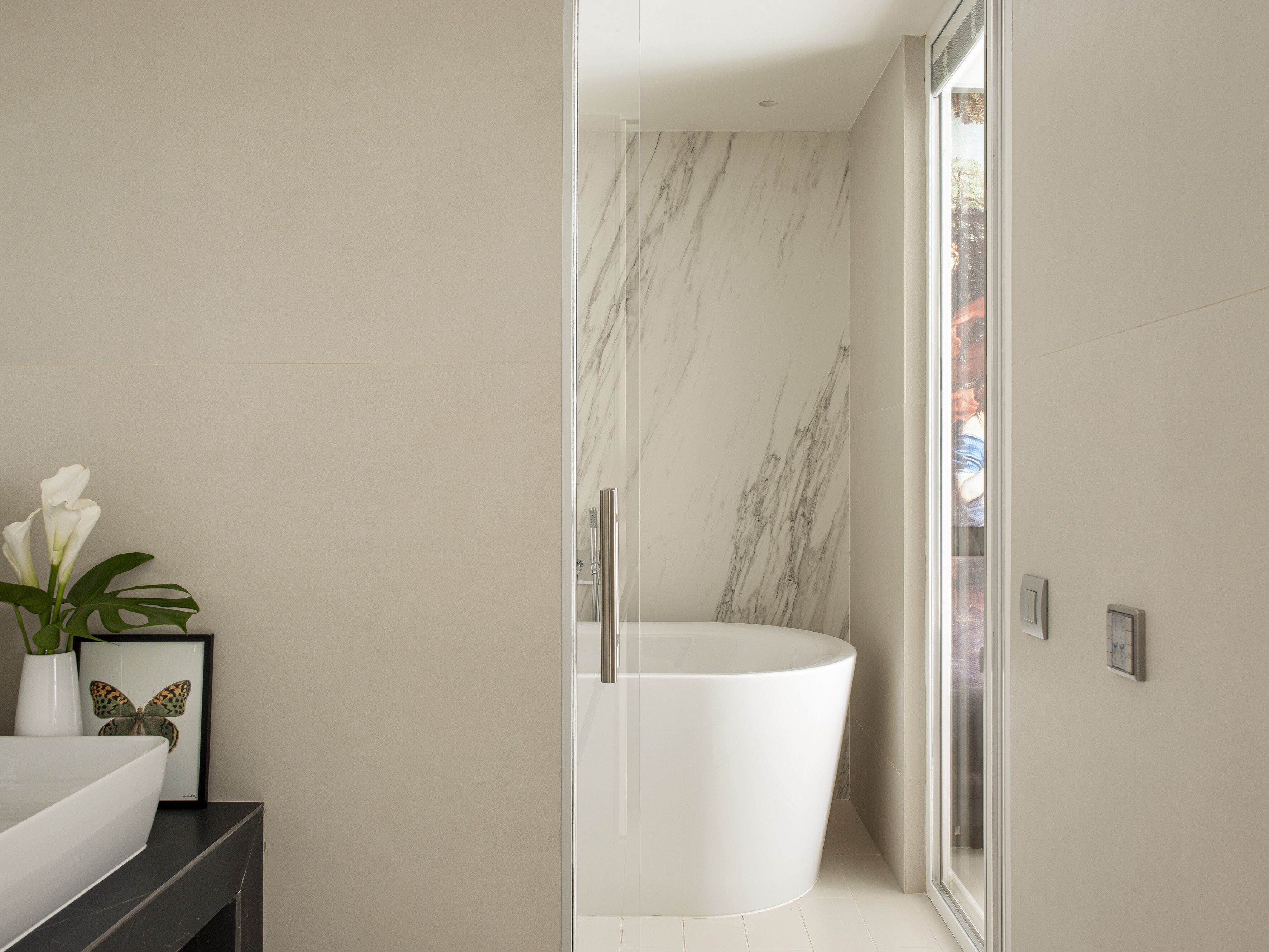 A modern bathroom with a marble wall, a white bathtub, and a vanity with a white sink and a vase with flowers.