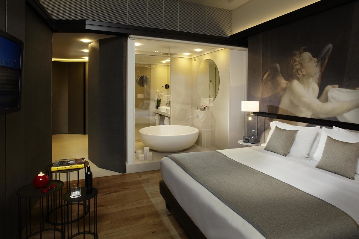 Una habitación de hotel moderna con una cama grande, un lavamanos blanco redondo, una mesita con una vela y un cuadro grande en la pared.