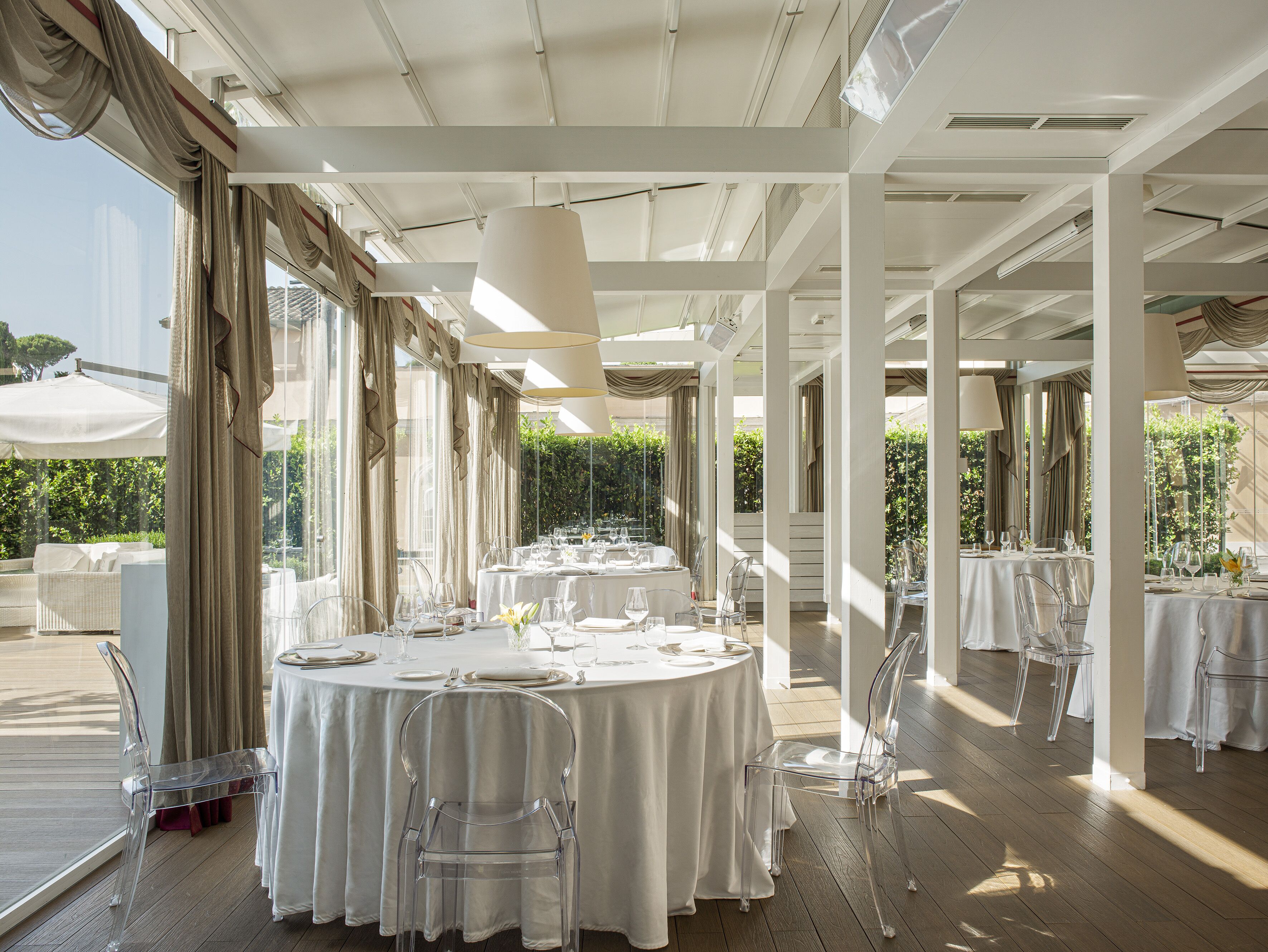 A restaurant with round tables, clear chairs, and white tablecloths.