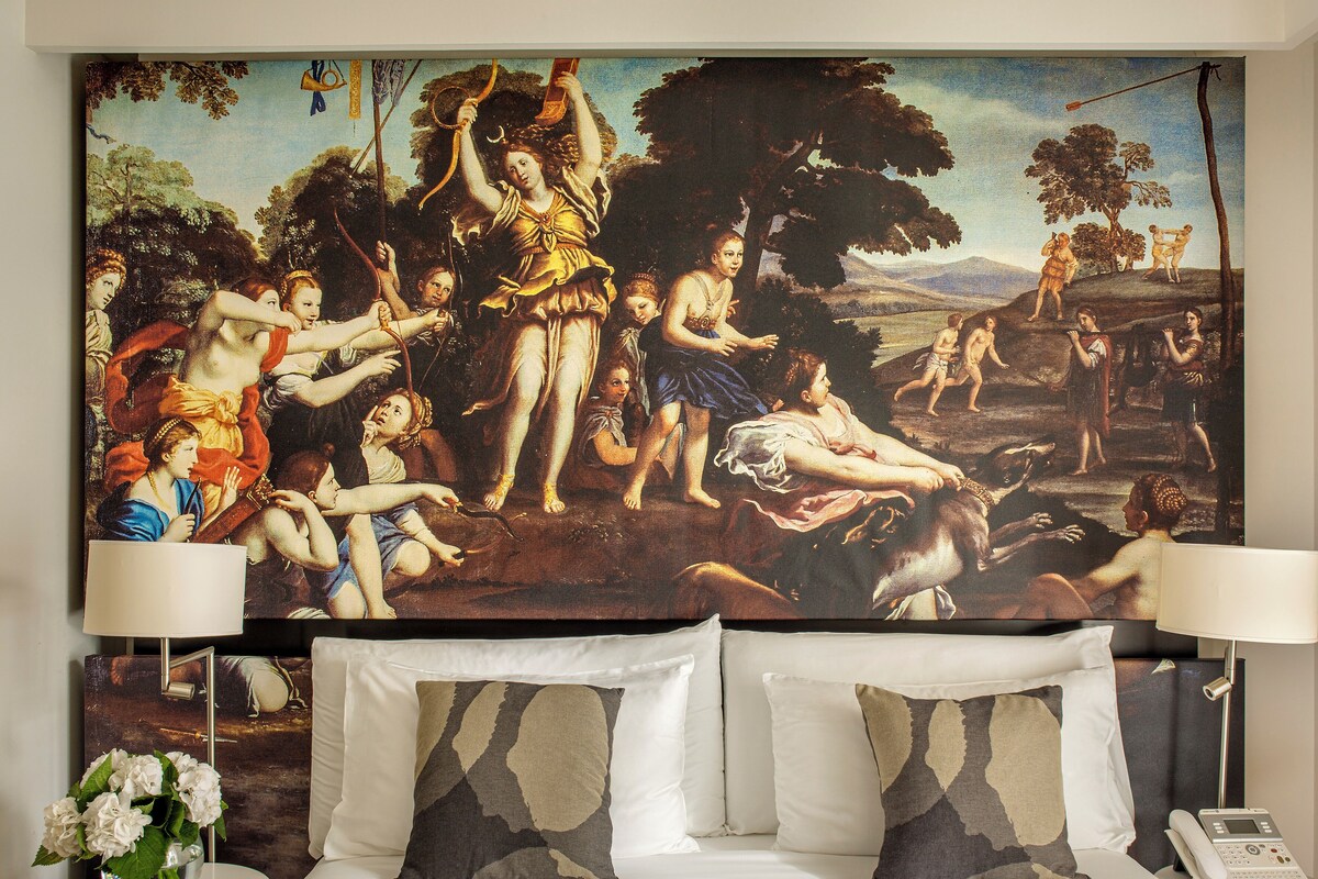 Habitación de hotel con un cuadro clásico grande en la pared, una cama con ropa blanca, dos almohadones decorativos, una mesita de noche con una lámpara y un jarrón con flores.