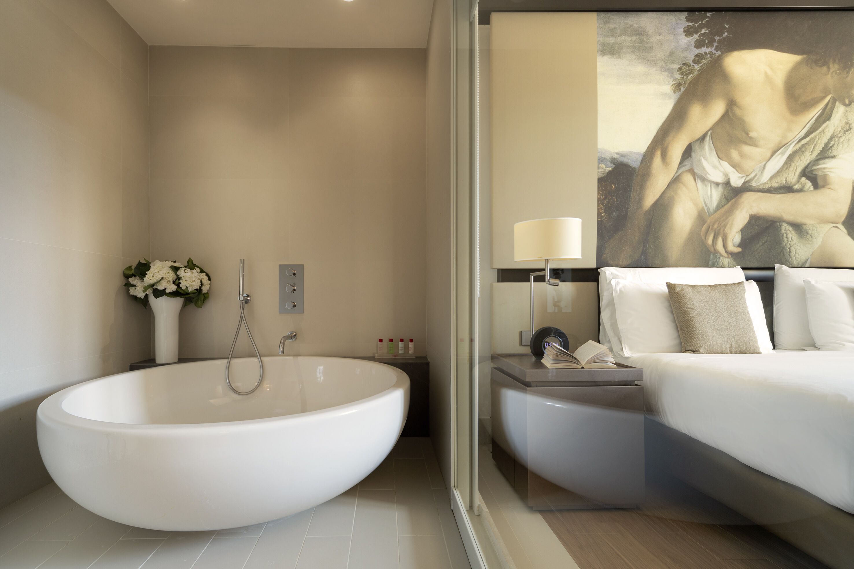 A modern bathroom with a large freestanding bathtub, a bedside table with a lamp, and a bed with white linens.