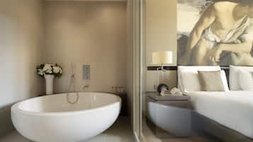 A modern bathroom with a large freestanding bathtub, a bedside table with a lamp, and a bed with white linens.