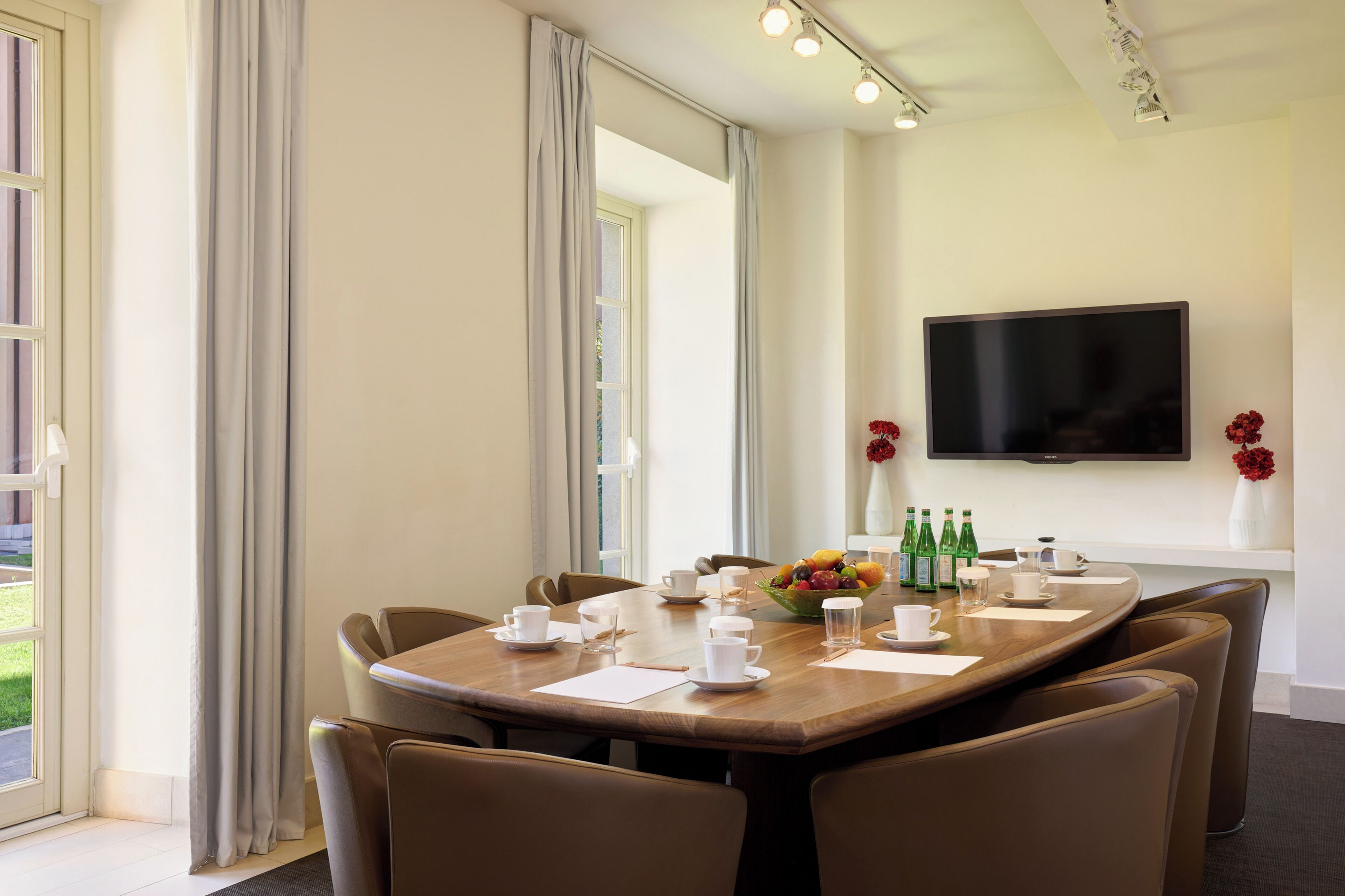 Una sala de conferencias con una mesa larga, sillas, un televisor montado en la pared y una ventana con cortinas.