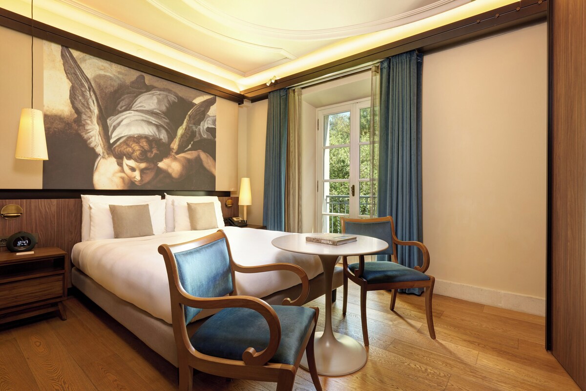 Habitación de hotel con una cama grande, dos sillas, una mesita, un cuadro en la pared y una ventana con cortinas.