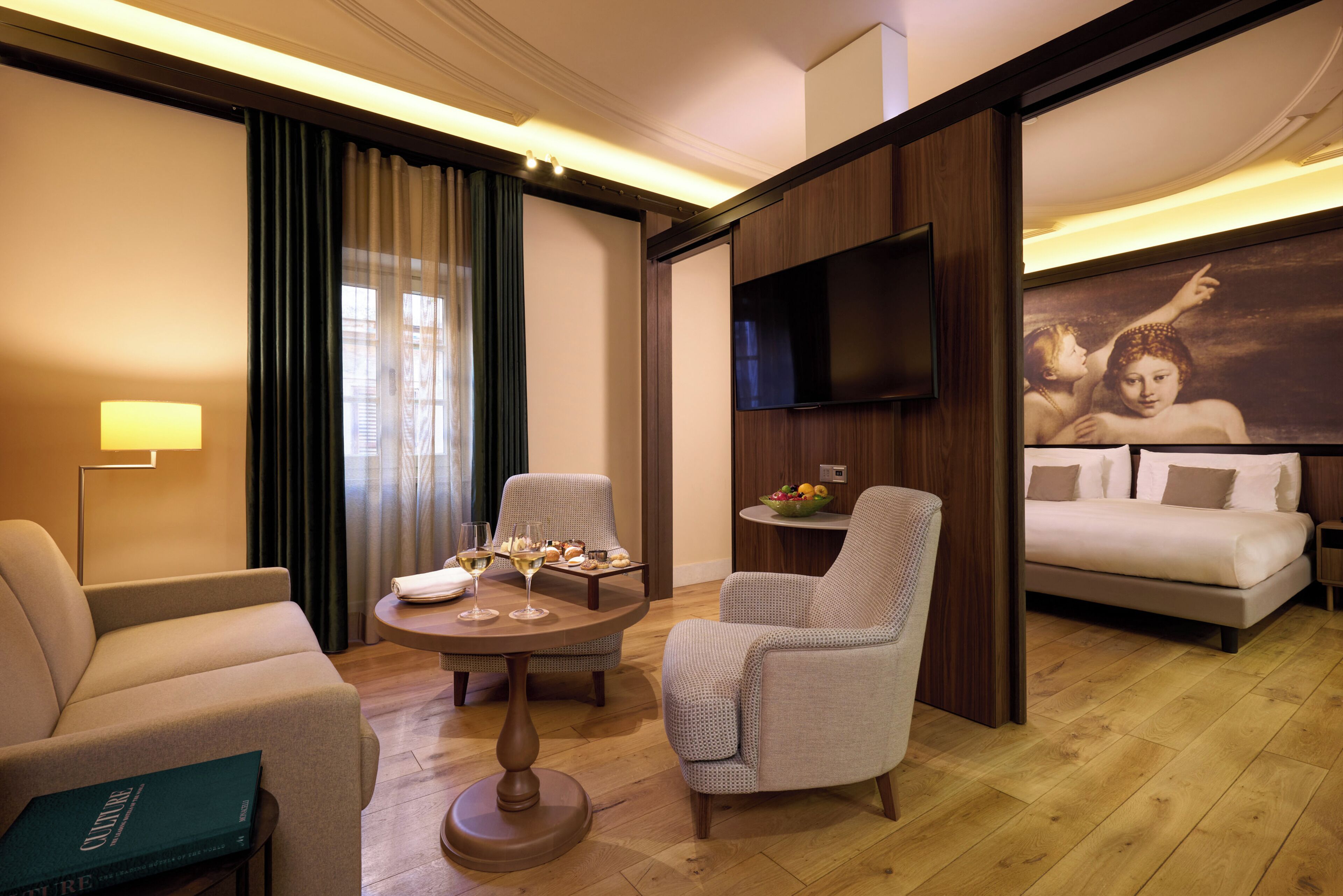 A modern hotel room with a sofa, armchairs, a coffee table, a flat-screen TV, a bed, and a large painting on the wall.