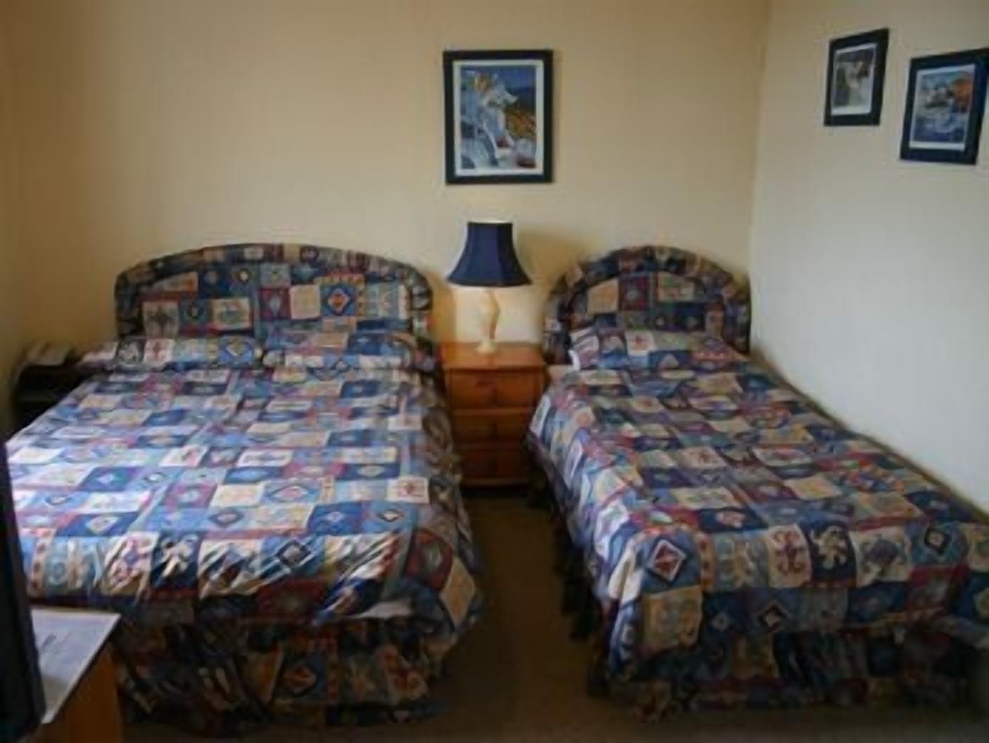 Une chambre avec deux lits, chacun recouvert d’une couvre-lit à motifs, une table de chevet avec une lampe et un tableau encadré au mur.