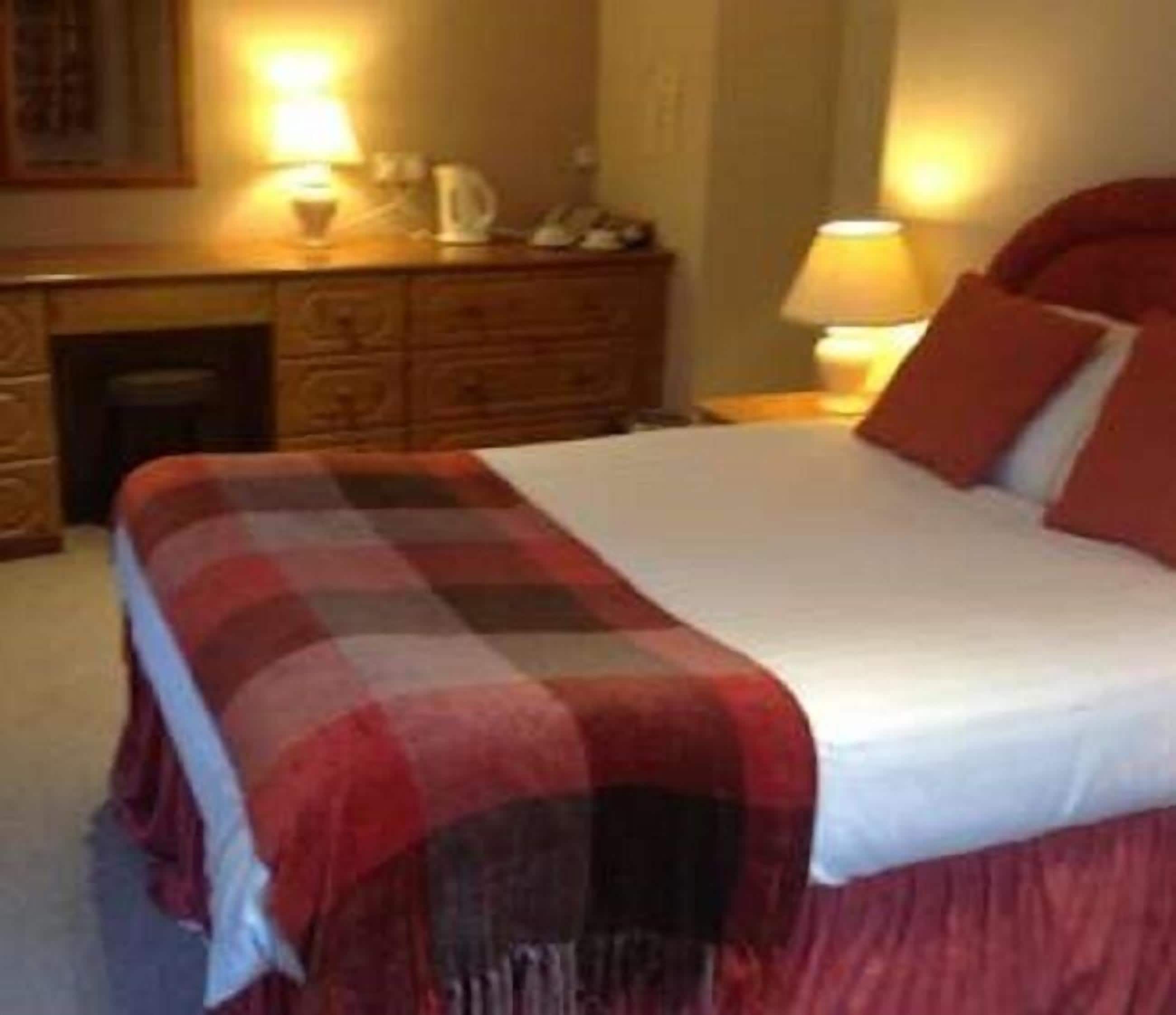 Un lit bien fait, recouvert d’une couverture à carreaux, une commode en bois avec une lampe, et une table de chevet avec une lampe.