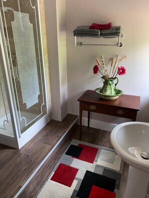 Een badkamer met een douche, een wastafel, een klein tafeltje met een bloemenvaas, een vloerkleed en een handdoekhouder.