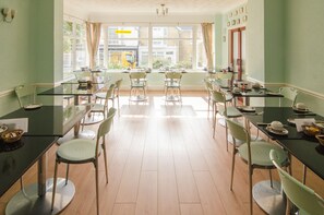 A dining area with multiple tables and chairs, large windows with curtains, and a view of the street outside. - The Braemar (Shanklin)