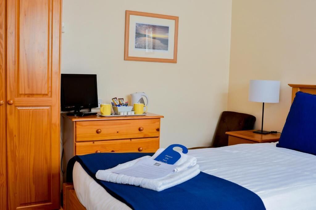 Ein Hotelzimmer mit einem Bett, einem Holzschrank, einem Schreibtisch mit Lampe, einem Stuhl und einem Fernseher auf einem Schrank.