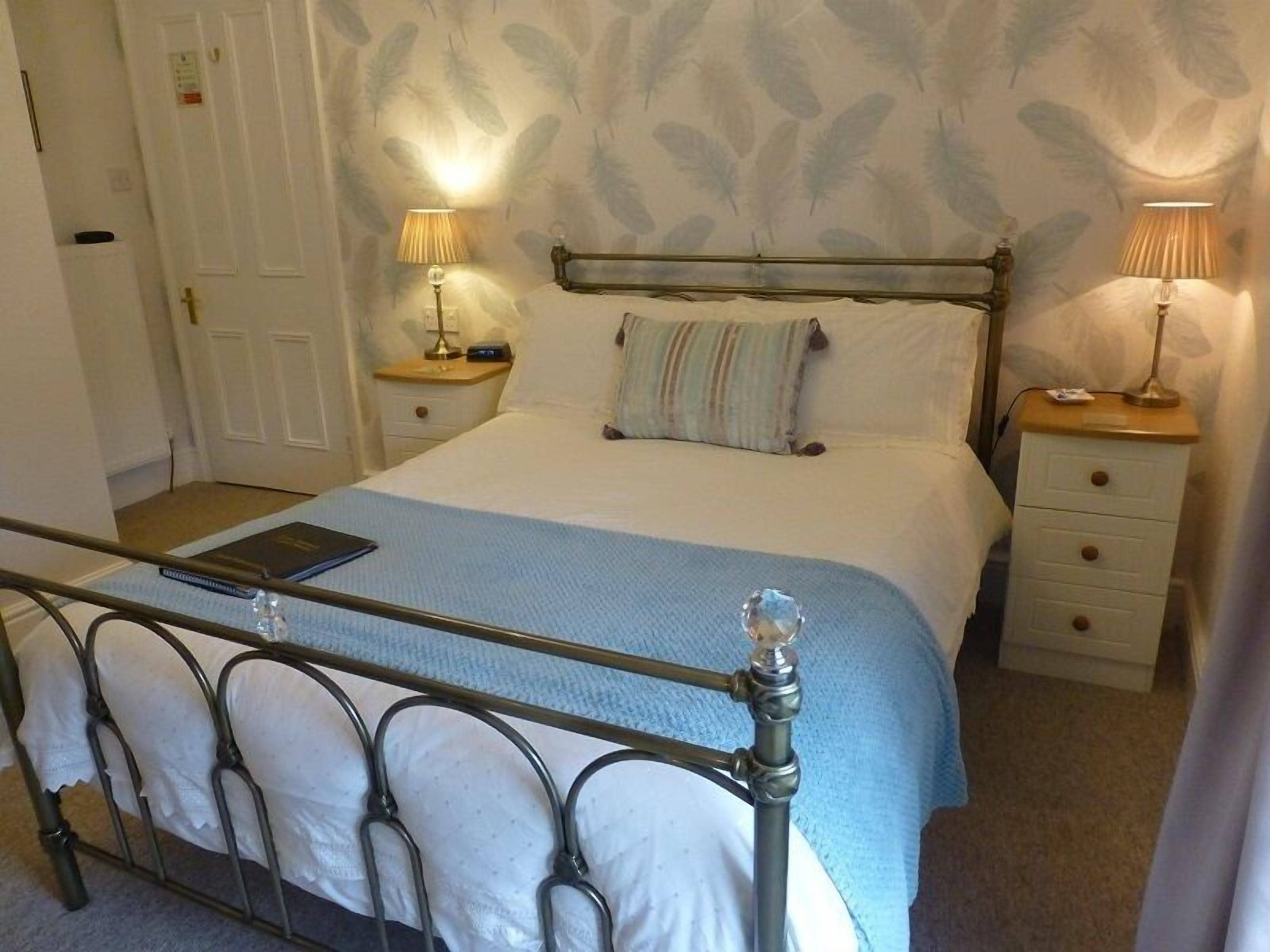 A bedroom with a metal bed frame, a bedside table with a lamp, a nightstand, a door, and a patterned wallpaper.