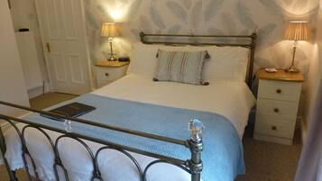 Un dormitorio con estructura de cama metálica, una mesita de noche con lámpara, una mesita auxiliar, una puerta y un papel tapiz estampado.