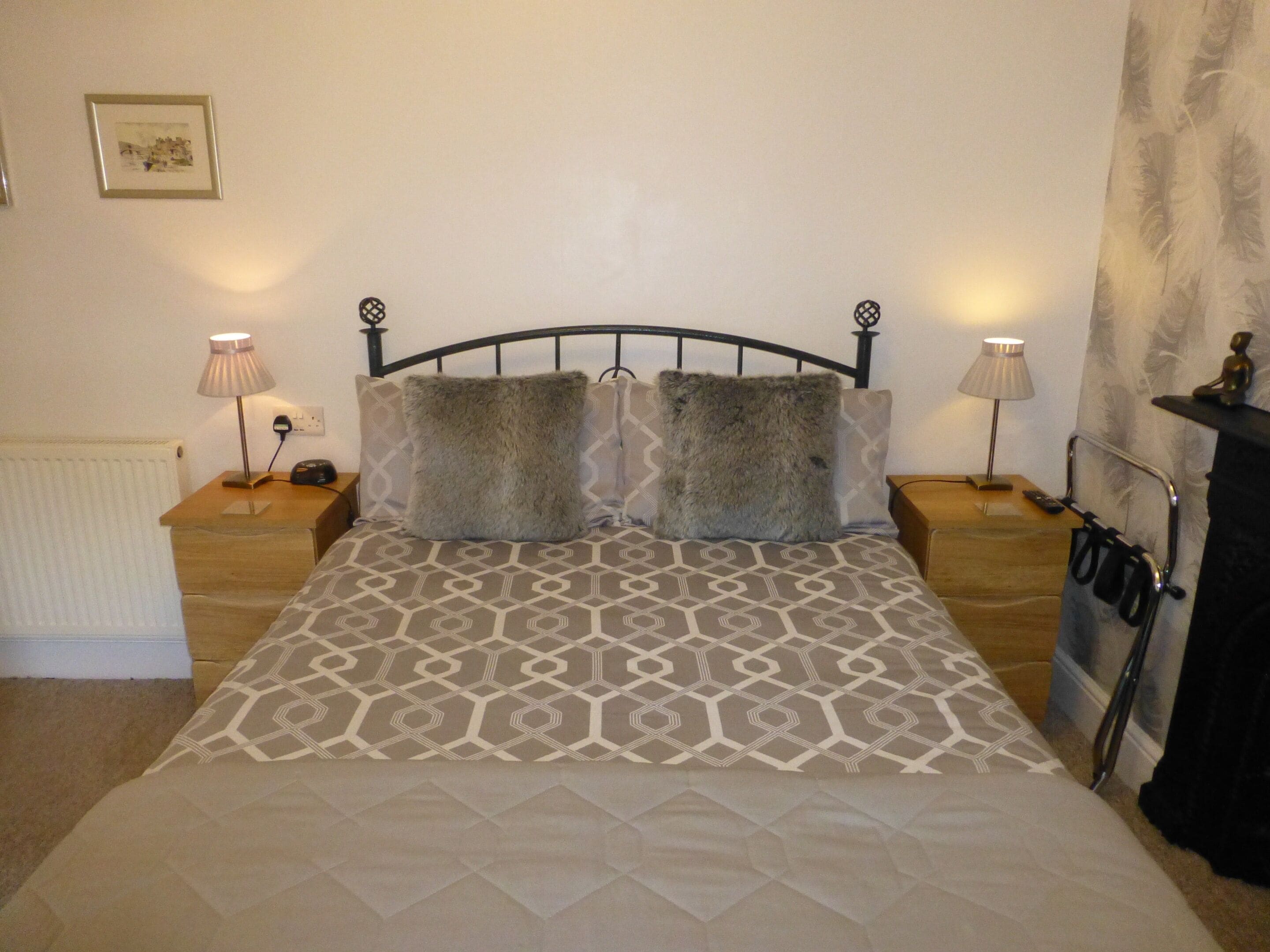 A neatly made bed with a patterned bedspread, two fluffy pillows, and a headboard. There are two bedside lamps, a radiator, and a framed picture on the wall.