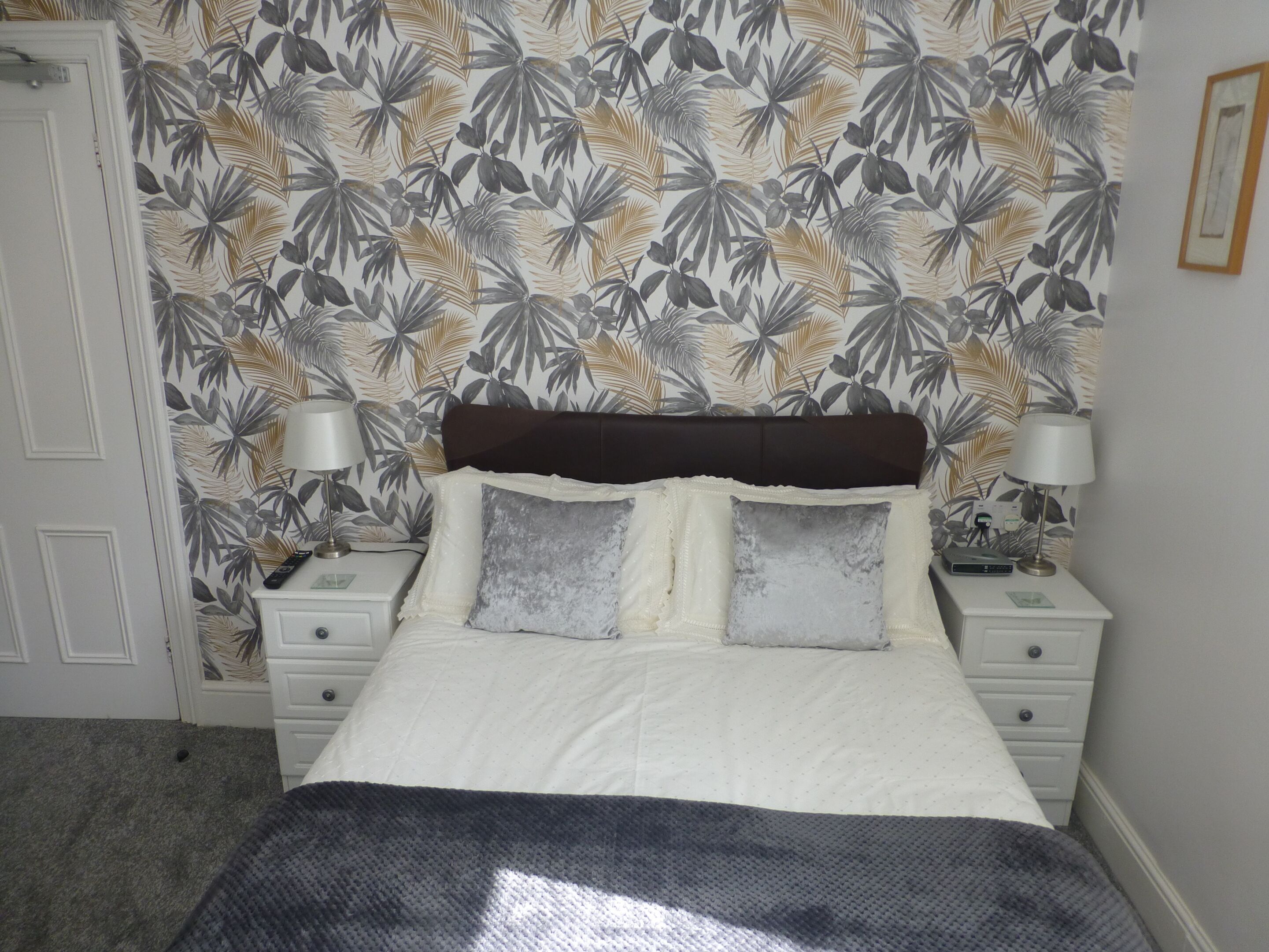 A bedroom with a bed, two bedside tables, a nightstand, a lamp, and a wall with a tropical leaf pattern.