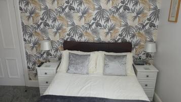 A bedroom with a bed, two bedside tables, a nightstand, a lamp, and a wall with a tropical leaf pattern.