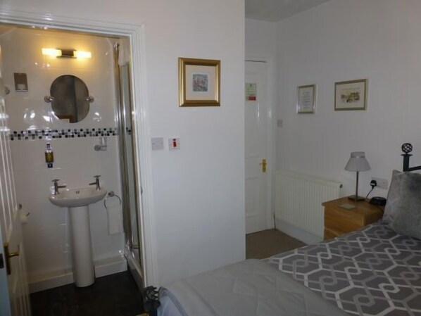 A bedroom with a bed, a bedside table with a lamp, a wall-mounted picture frame, and a bathroom with a sink and mirror. - Bryn Derwen Guest House (Conwy)