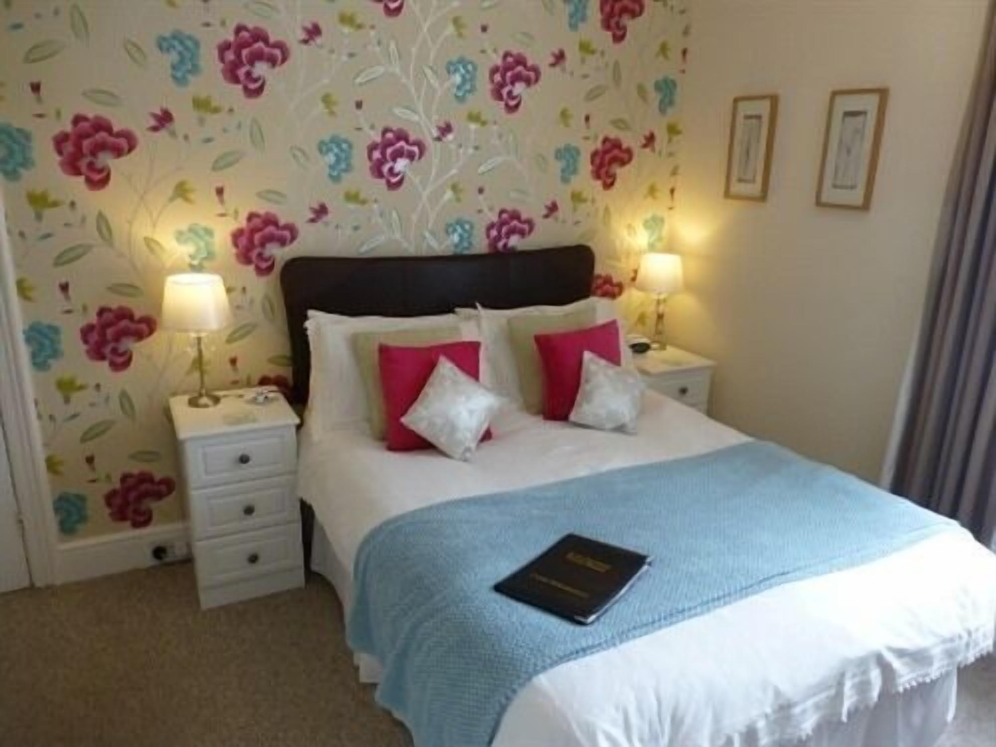A neatly made bed with a blue blanket and pillows, two bedside lamps, and a floral wallpaper pattern.