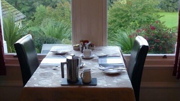 A dining table set for two with a view of a lush garden through a window. - Bryn Derwen Guest House (Conwy)