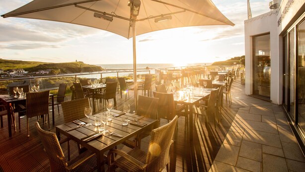 An outdoor dining area with tables and chairs, a large umbrella, and a view of the sea.