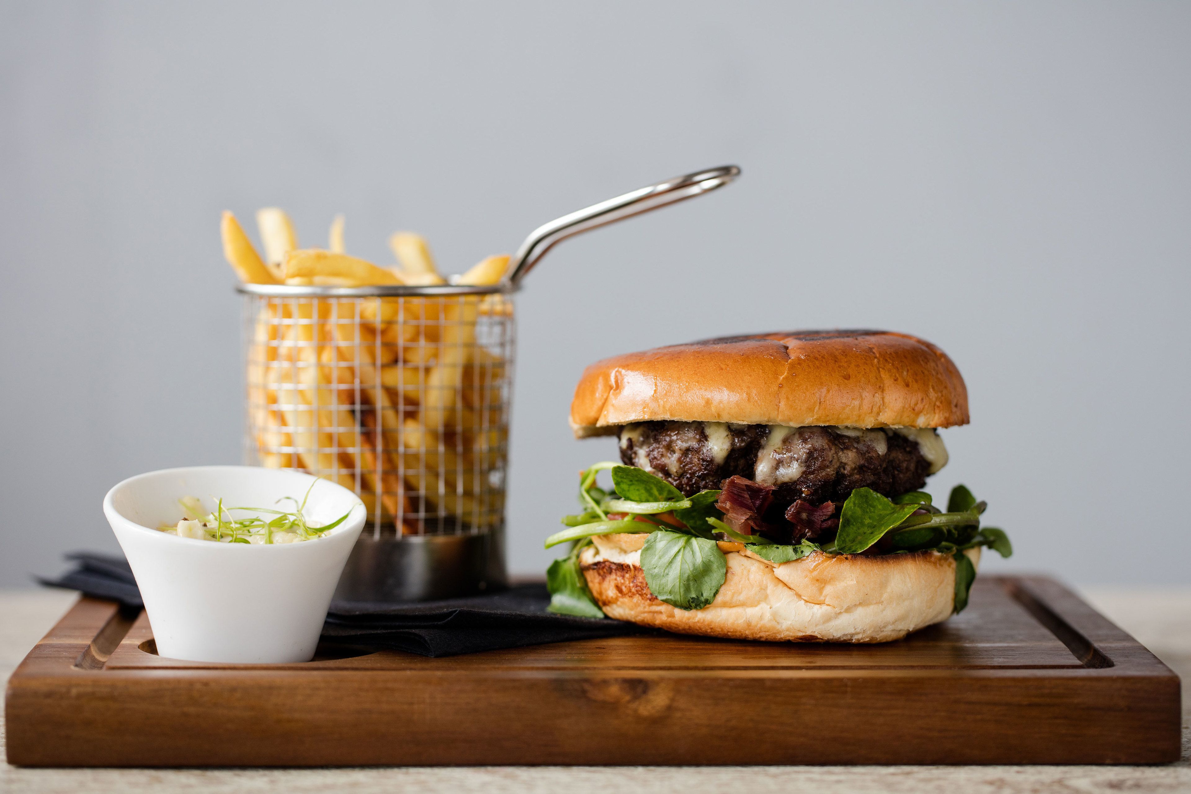 A burger with fries and a small bowl of greens on a wooden board.