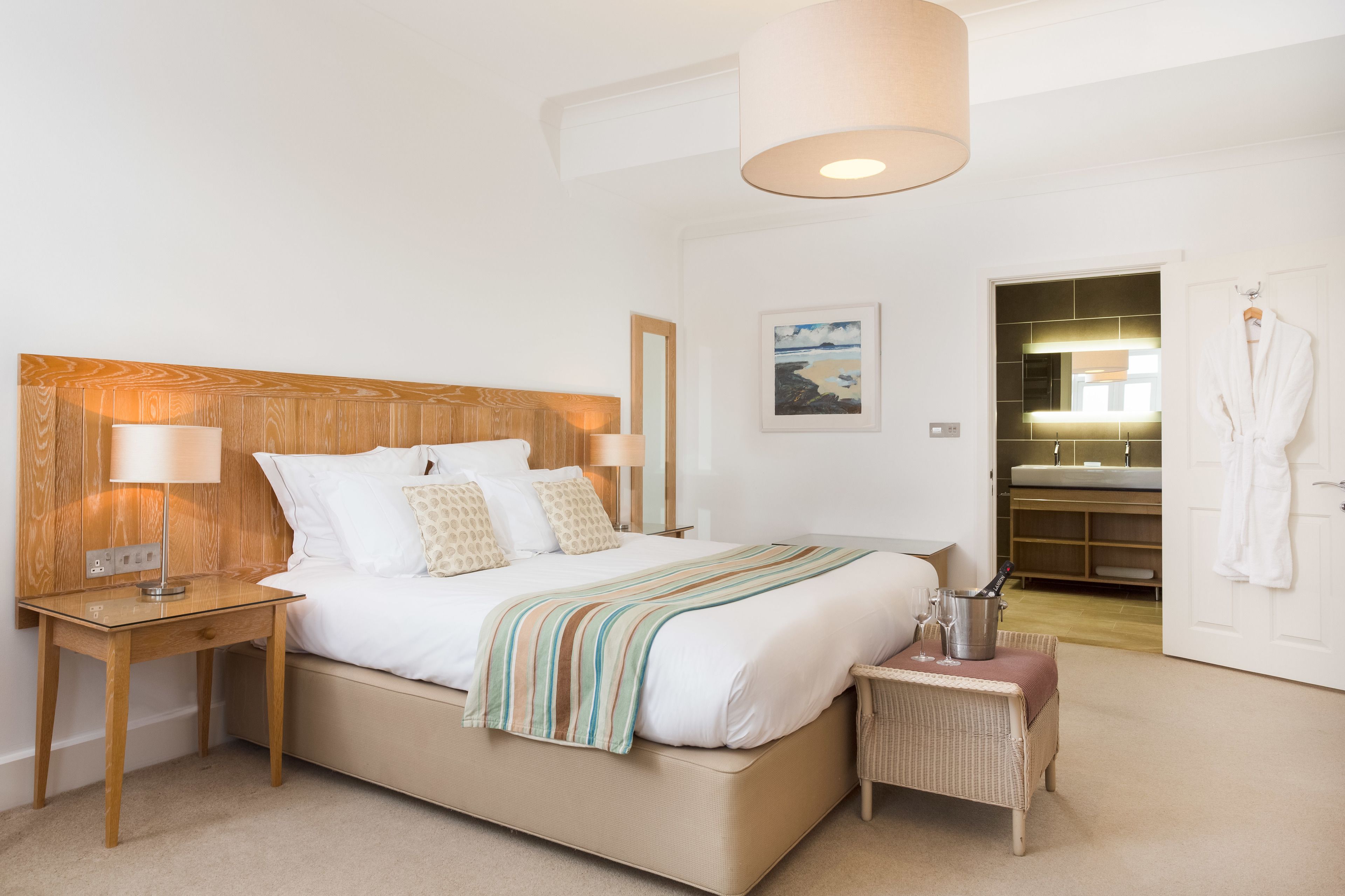 A bedroom with a large bed, bedside tables, a nightstand, a small table with glasses, a hanging robe, and a bathroom visible through an open door.
