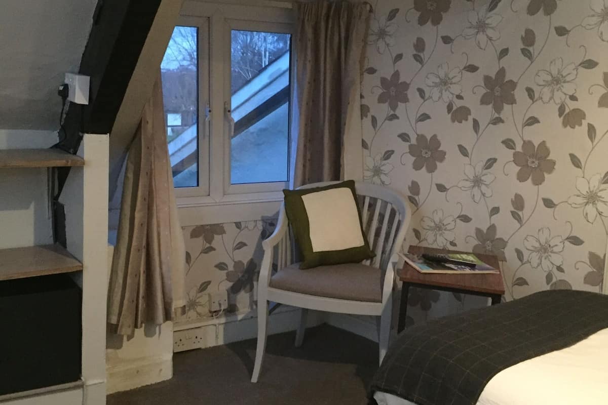 A bedroom with a sloped ceiling, a window with curtains, a chair with a pillow, a bedside table with a book, and a bed with a blanket.
