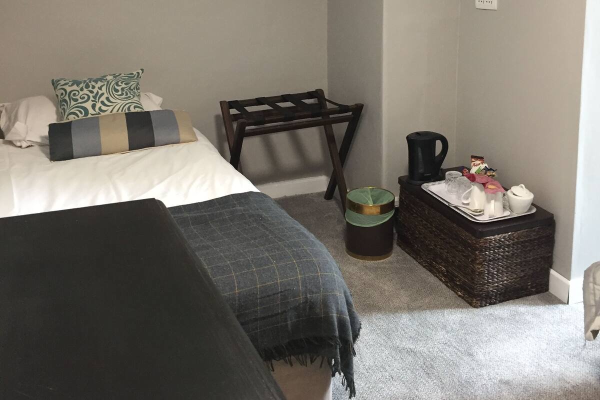 A bedroom with a bed, a chair, a small table with a kettle, and a basket.