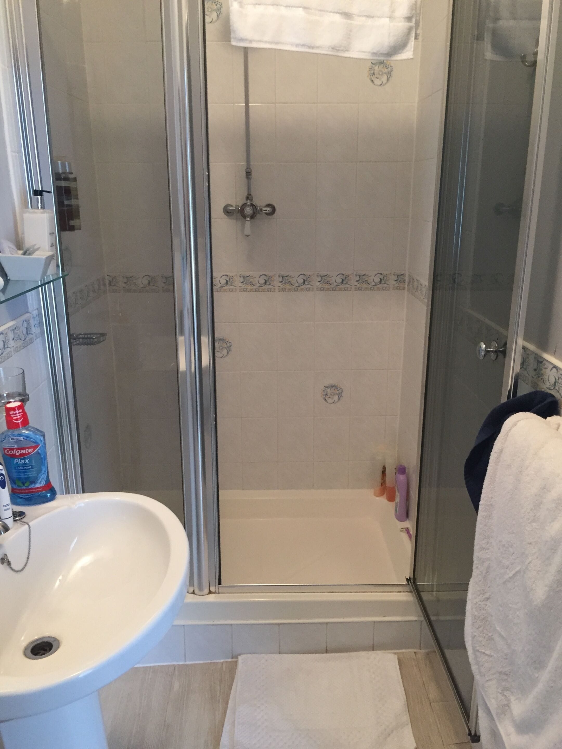 Et badeværelse med en brusekabine med glasvægge, en hvid vask og en glashylde med toiletartikler.