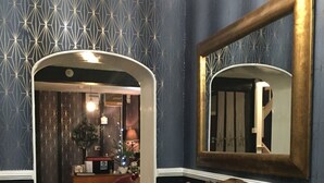 A room with a geometric patterned wallpaper, a large mirror, a wooden table with toiletries, and a red armchair. - Castle Lodge Hotel (Ross-on-Wye)