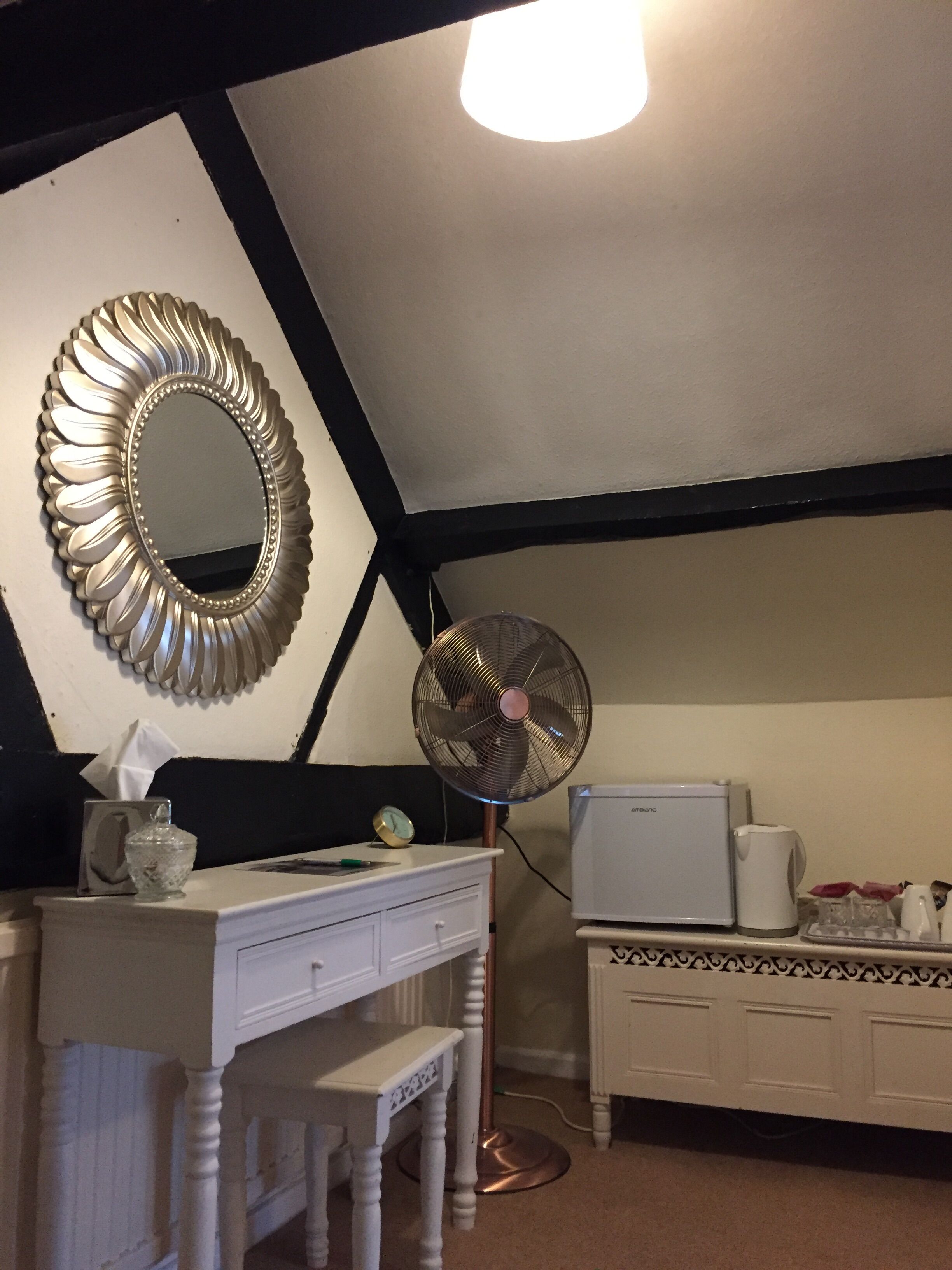 A bedroom with a white dresser, a mirror with a decorative frame, a standing fan, a small table with a tissue box, a clock, and a white cabinet with patterned trim.