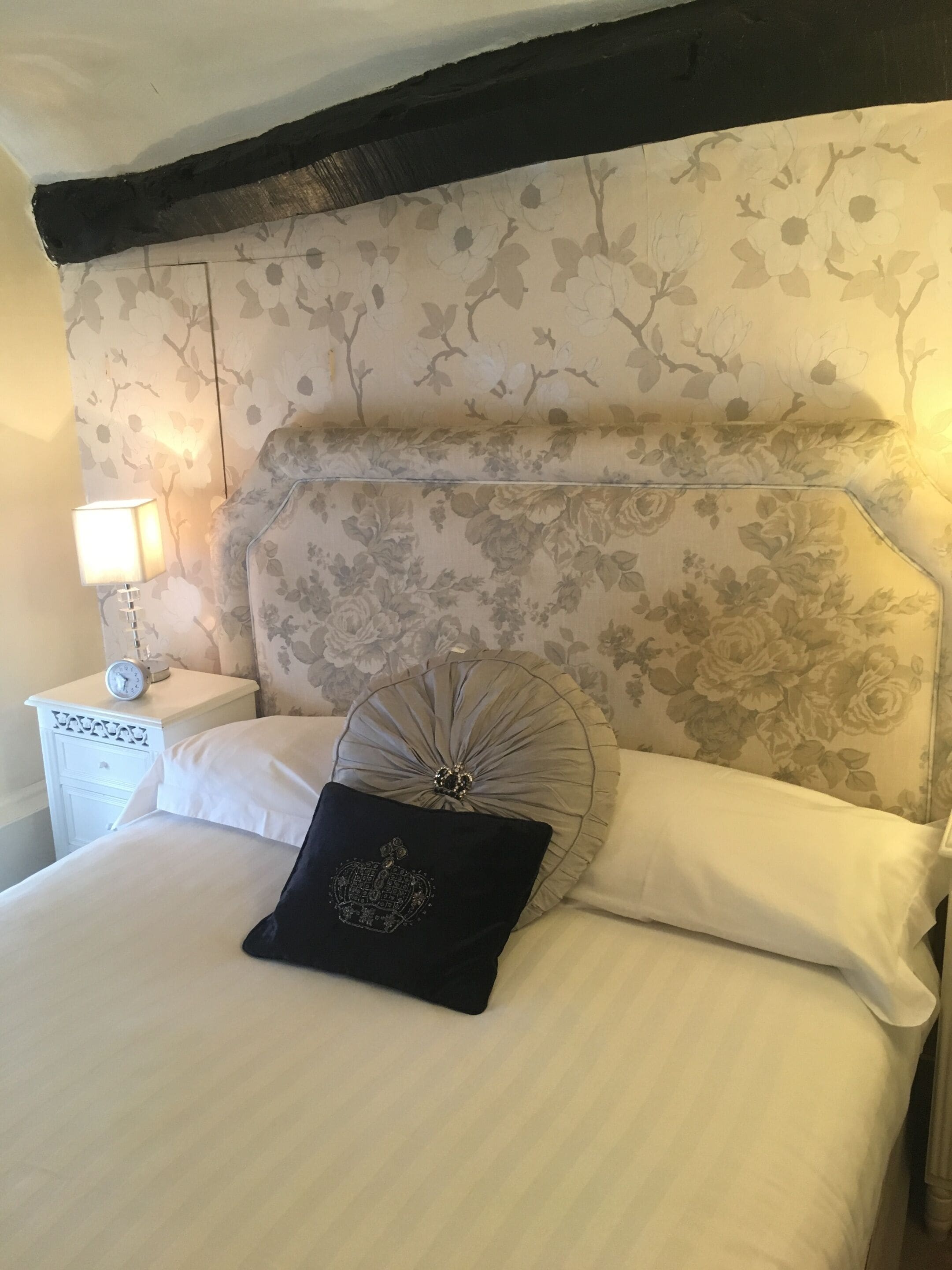 Un dormitorio con cabecera estampada, una mesita de noche blanca con un reloj y una cama con ropa de cama blanca y almohadas decorativas.