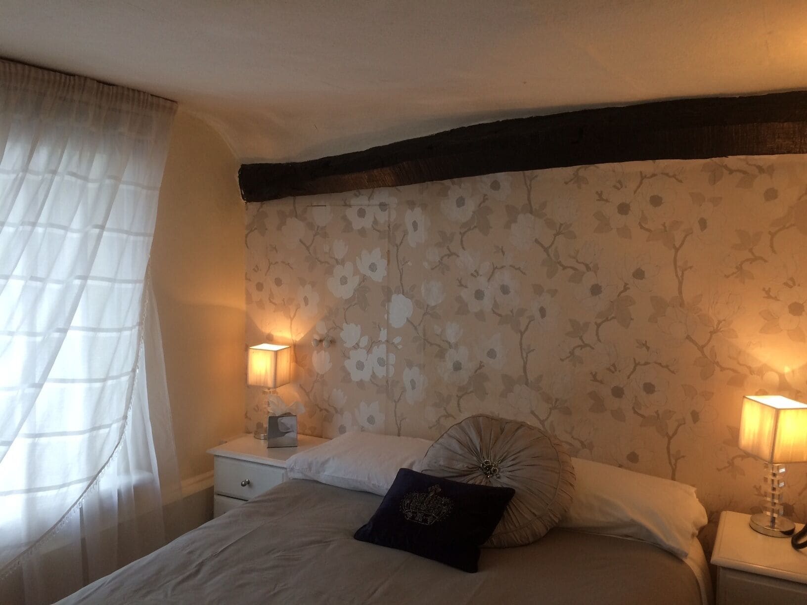Une chambre à coucher avec un papier peint à motifs floraux, un lit avec deux oreillers, deux lampes de chevet et une table de chevet sur laquelle se trouvent une boîte de mouchoirs et un petit objet décoratif.