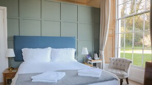 A neatly made bed with white linens, a grey throw blanket, and folded towels. There is a small bedside table with a lamp and a decorative item. A chair is placed next to the window, and a large window offers a view of greenery outside. - Laston House (Ilfracombe)