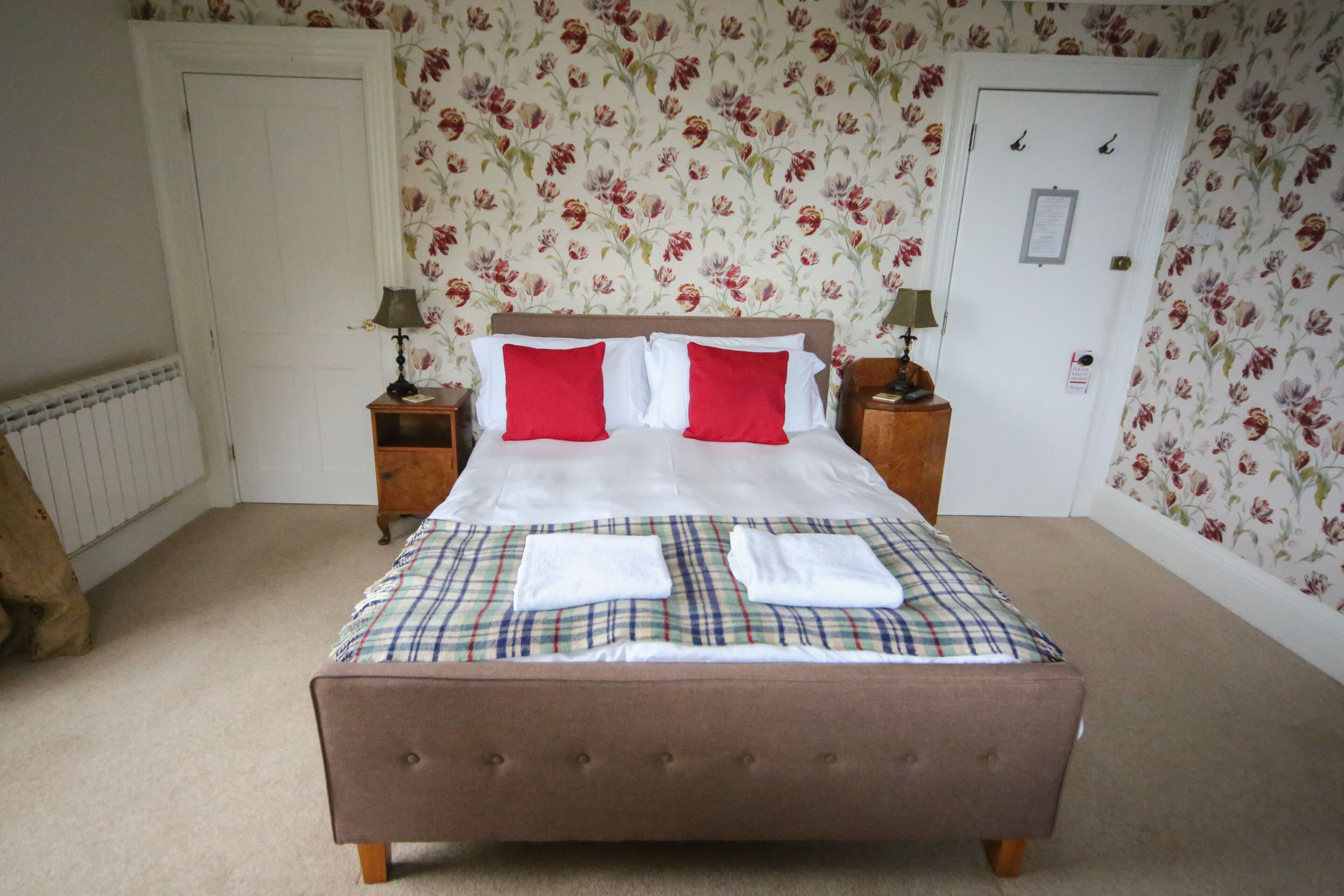 A neatly made bed with a plaid bedspread and red pillows, flanked by nightstands with lamps, in a room with floral wallpaper and a white door.