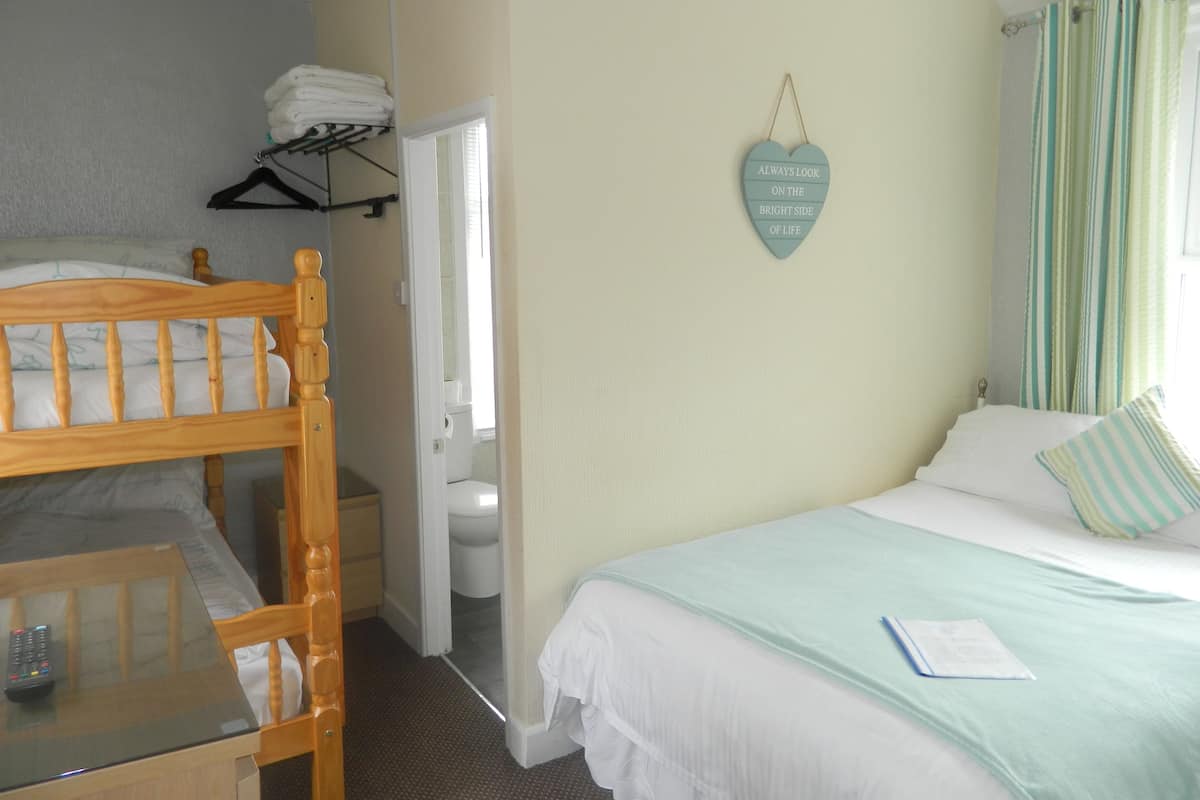 A bedroom with a bunk bed, a bedside table with a remote control, a bed with a light blue bedspread and a pillow, and a wall-mounted heart-shaped sign.