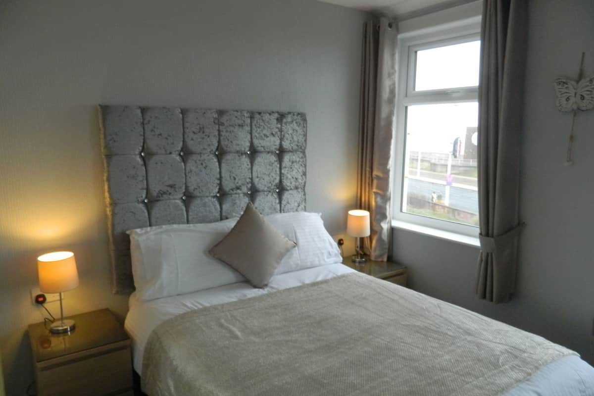 A neatly made bed with a tufted headboard, two bedside lamps, and a window with curtains.